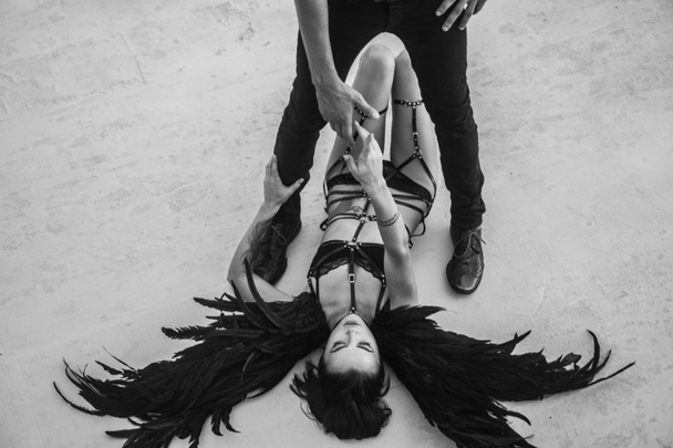 Foto en blanco y negro de la escena romántica con la silueta del hombre de pie por encima de la sensual mujer ángel con lencería, cinturones de cuero y tacones altos tirados en el suelo
 - Foto, imagen