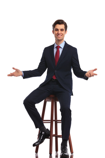 молодий щасливий бізнесмен сидить на стільці і вітає вас
 - Фото, зображення