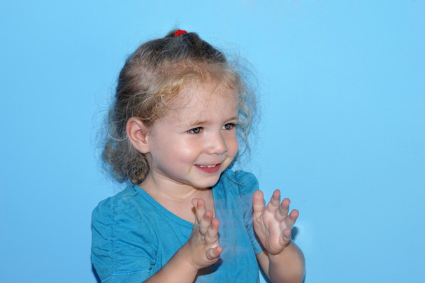 Smiling little girl with blue T-shirt claps her hands on blue background close-up - Photo, Image