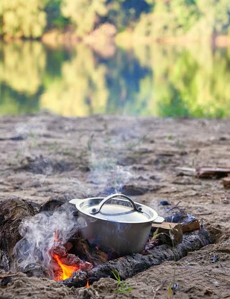 cooking on fire at picnic, food prepared in kettle on wood, trees reflected in river, healthy vegetarian food - Photo, Image
