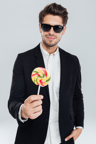 Businessman in sunglasses standing with hand in pocket holding lollipop - Zdjęcie, obraz