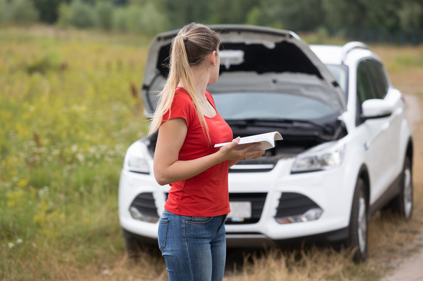 Mujer joven mirando su coche roto y leyendo manual del propietario
 - Foto, Imagen