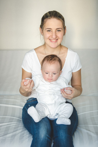 Mooie lachende moeder zittend op bed en houden haar babyjongen - Foto, afbeelding