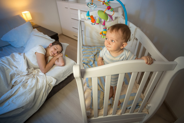 Портрет ребенка, стоящего в кроватке и глядя на усталую мать й
 - Фото, изображение