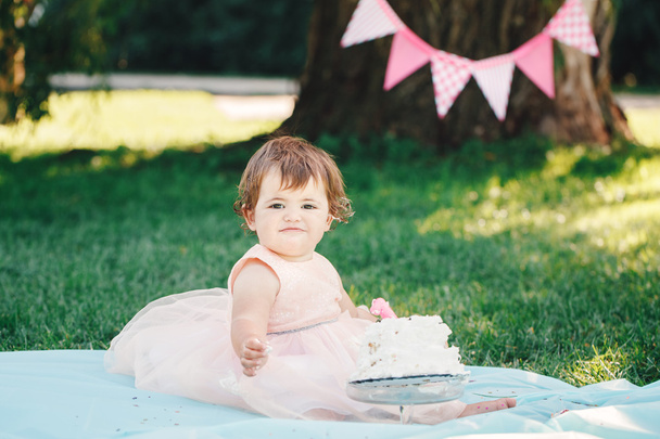 Retrato de bonito adorável menina branca com olhos castanhos escuros em vestido de tutu rosa comemorando seu primeiro aniversário com bolo gourmet olhando na câmera do lado de fora no parque, quebra de bolo
 - Foto, Imagem