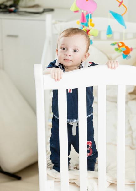 Cute 9 month old boy standing in white wooden crib - Foto, afbeelding