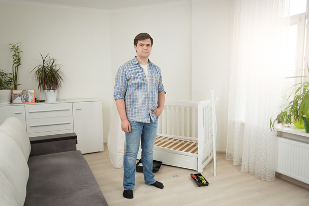 Handsome man standing at disassembled baby crib at bedroom - Фото, изображение