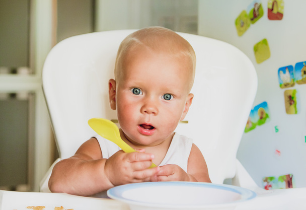 Cute baby  with a spoon in the high chair on the kitchen - Photo, Image