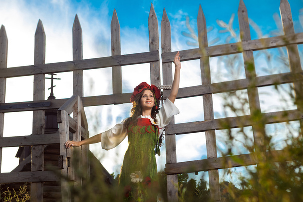 Молодая женщина в русском традиционном платье стоит рядом с деревянным
 - Фото, изображение