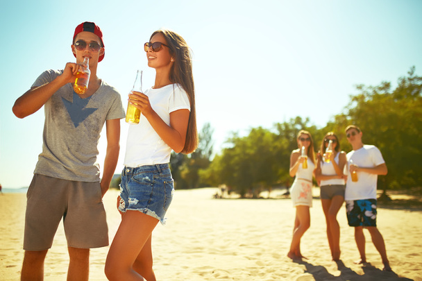 Lächelndes junges Paar, das Gläser mit Bier klappert, während im Hintergrund gegrillt wird. Die Menschen genießen die Sommerzeit. Spaß am Strand. Tolle Sommerstimmung. sonniger Tag - Foto, Bild