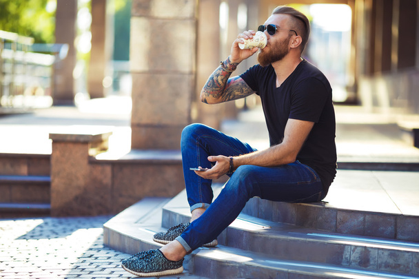 Handsome young man holding mobile phone and drinking coffee while sitting in the rest area of the office.  - Foto, imagen
