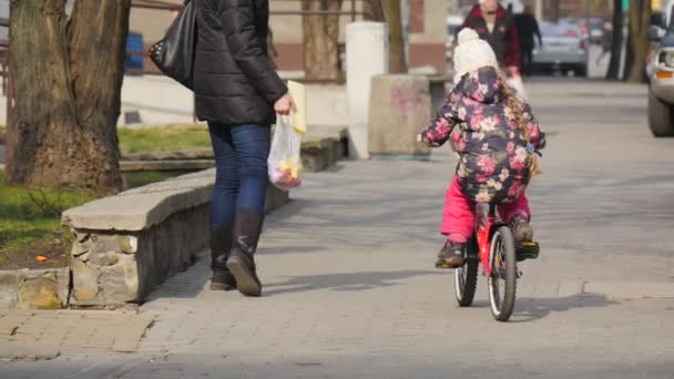 Family Mum and Daughter on a City Street People Parked Cars Traffic Cityscape Little Girl is Riding a Bicycle Woman is Walking Along the Kid on a Sidewalk - Filmati, video