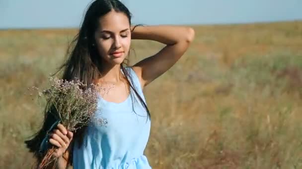 Jeune belle fille dans une robe bleue aime les fleurs steppe. Une jeune femme dans un pré. Steppe. Pré ensoleillé fleurissant avec de hautes épis d'herbe épaisses oreilles et fleurs
 - Séquence, vidéo