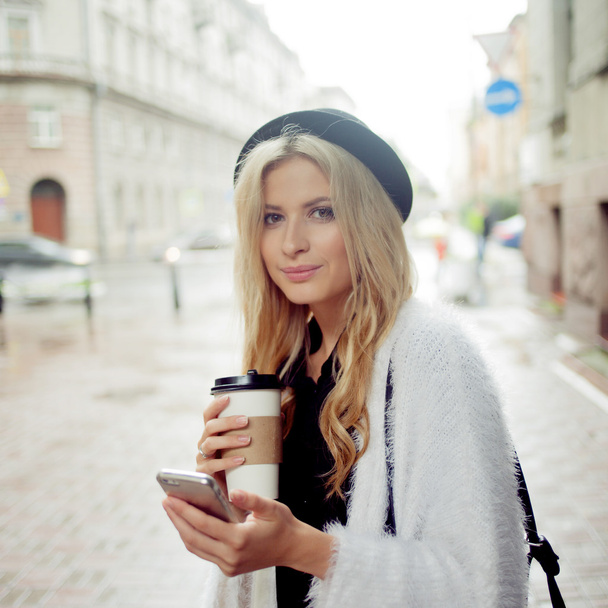 Веселая женщина на улице пьет утренний кофе и пользуется смартфоном
. - Фото, изображение