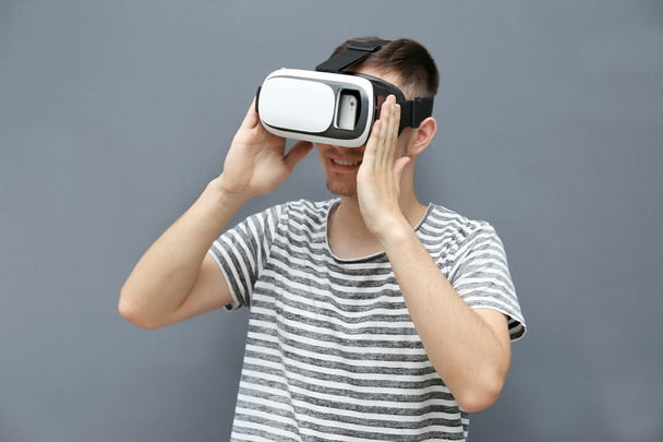 Young man in striped T-shirt wearing virtual reality glasses on grey background - Photo, Image