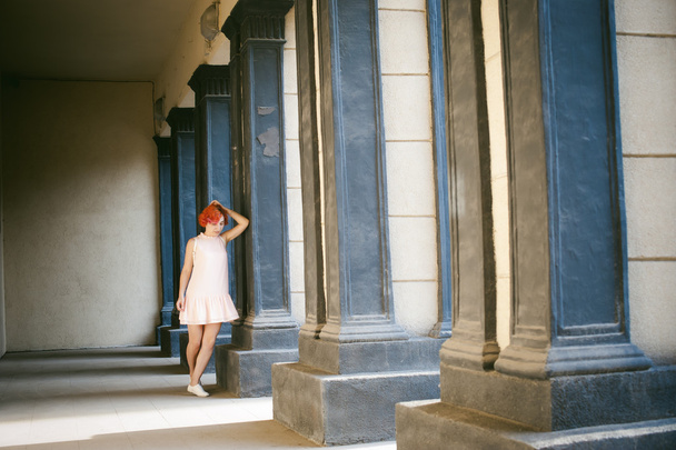 girl in a pale pink dress with dyed red hair, a white backpack in hand, walking between light and shadow on the columns sunny summer day - 写真・画像