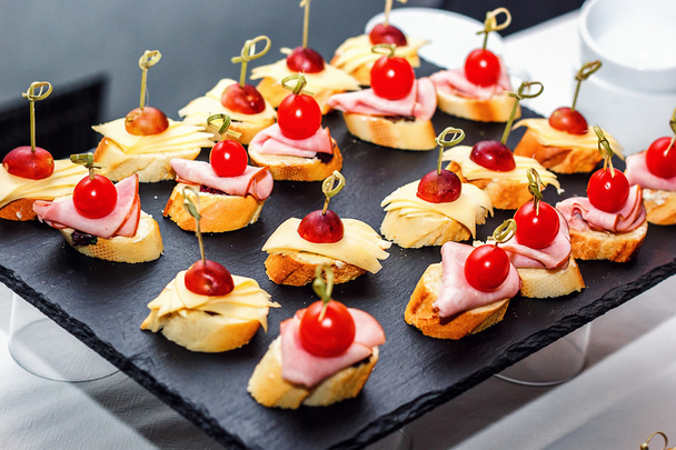 Small crostini with grilled baguette, cherry tomatoes, ham slices, cheese and fresh grapes on black background. Sandwiches or assorted canapes on a dark board, top view. - Photo, Image