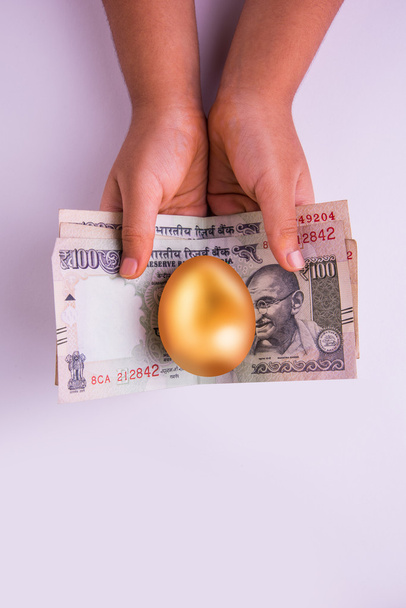 closeup of small girl holding indian currency notes or hundred rupee notes and golden egg over it - Photo, Image