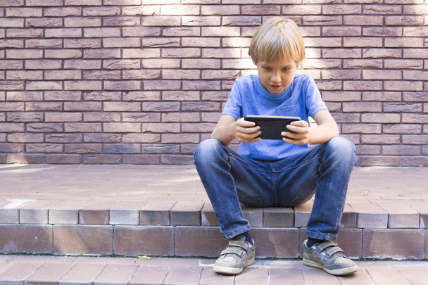 Child with mobile phone sitting outdoors. Boy looks at the screen, use application, plays. Sunny day. Brick wall building background. people, technology, leisure concept - Фото, зображення