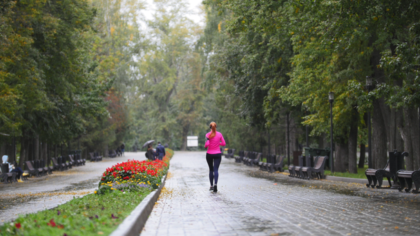 Juoksija nuori nainen juoksee syksyllä puistossa treenaamassa ulkona sateella, vaaleanpunainen harjoituspuku, peruutusnäkymä
 - Materiaali, video