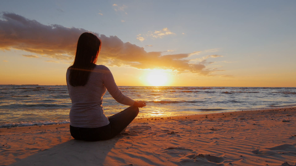 Χωρίς στρες. Ήρεμη νεαρή γυναίκα κάθεται σε μια θέση λωτού στην παραλία στο ηλιοβασίλεμα - Πλάνα, βίντεο