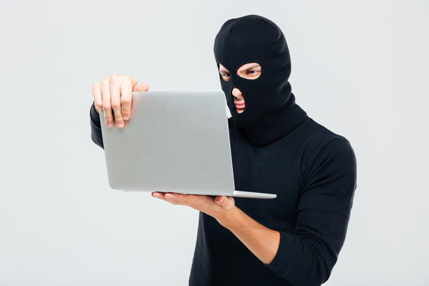 Criminal young man in balaclava standing and using laptop - Foto, Bild