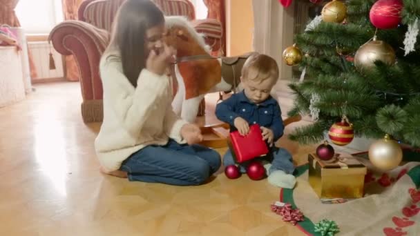 Cute baby boy with elder sister sitting on floor under Christmas tree and opening boxes with gifts - Footage, Video