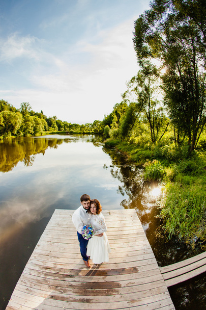 The bride and groom are standing on a wooden pier near the pond - Foto, imagen