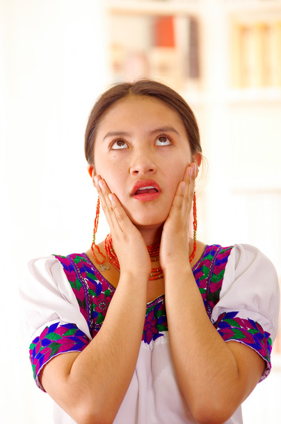 Beautiful young brunette woman wearing traditional andean white blouse with blue decorative edges, facing camera interactin frustrated, bright household background - Photo, image
