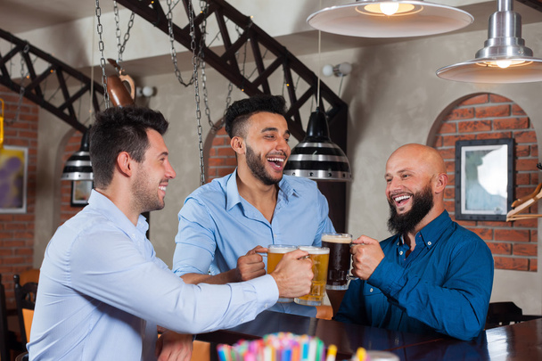 Three Man In Bar Clink Glasses Toasting, Drinking Beer Hold Mugs, Mix Race Cheerful Friends Wear Shirts - 写真・画像