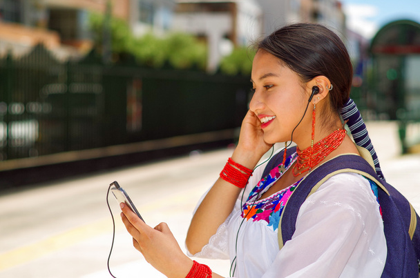 Pretty young woman wearing traditional andean blouse and blue backpack, waiting for bus at outdoors station platform, looking to mobile screen with headphones connected smiling happily - Photo, Image