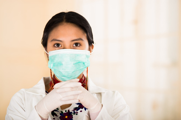 Young beautiful woman dressed in doctors coat and red necklace, face covered with facial mask looking into camera, egg white clinic background - Foto, Imagen