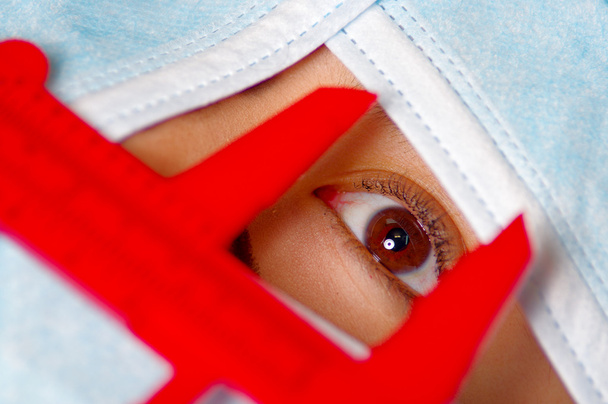 Closeup eye of woman peeking out from total facial cover, preparing for cosmetic surgery concept, doctor using red measure tool - Photo, image
