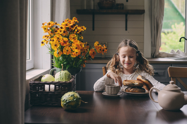 child girl having breakfast at home in autumn morning. Real life cozy modern interior in country house. Kid eating bagels and drinking tea.  - Photo, Image