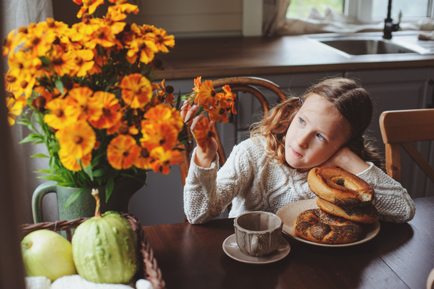 child girl having breakfast at home in autumn morning. Real life cozy modern interior in country house. Kid eating bagels and drinking tea.  - Foto, immagini
