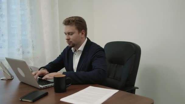 Young businessman working in his office in front of the laptop computer then he looks at his watch understands that he is late for a meeting takes his stuff and goes away - Footage, Video