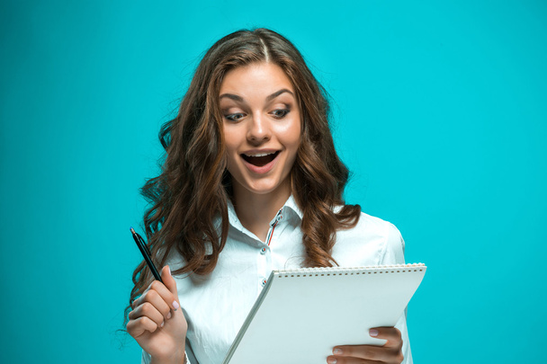Surprised young business woman with pen and tablet for notes on blue background - Fotó, kép