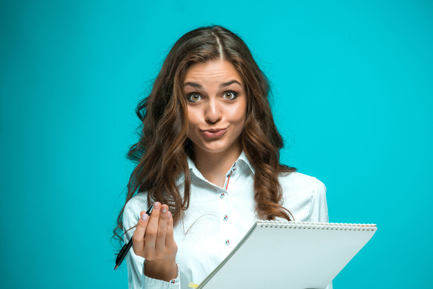 Surprised young business woman with pen and tablet for notes on blue background - Фото, изображение