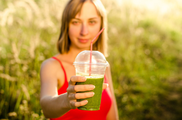  πρόσωπο από μια χαμογελαστή γυναίκα στα χέρια πράσινη ενέργεια και διατροφική smoothie κοκτέιλ στο φόντο θολή φύση - Φωτογραφία, εικόνα