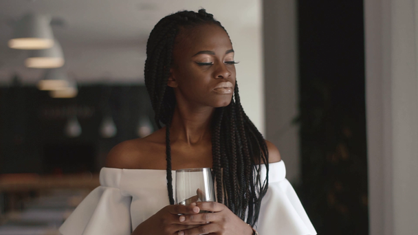 Young african american female fashion model holding empty wineglass in her hand, standing near illuminated window with white curtains - Footage, Video