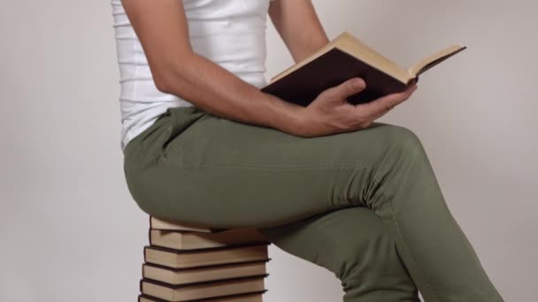 Man in groene broek leest een boek, zittend op een grote stapel boeken. Grijze achtergrond. 4k schot - Video