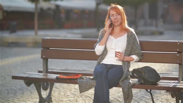 Mooie vrouw met rood haar praten over de mobiele telefoon zittend op een bankje in de straat met gebouwen in de achtergrond, meisje, drinken koffie en lachen - Video
