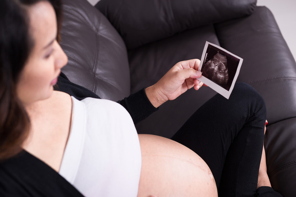Pregnant woman sitting on sofa, holding her child ultrasound pic - 写真・画像