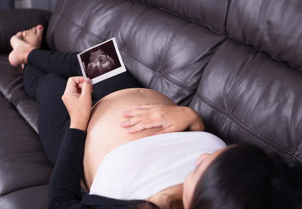 Pregnant woman resting on sofa, holding her child ultrasound pic - 写真・画像