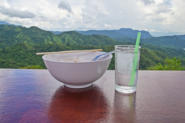 Drinking glass and bowl of noodles are placed on wooden floor. There is a beautiful mountain backdrop  - Photo, Image