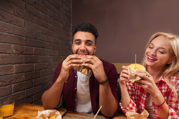Young Man And Woman Eating Fast Food Burgers Sitting At Wooden Table In Cafe - 写真・画像