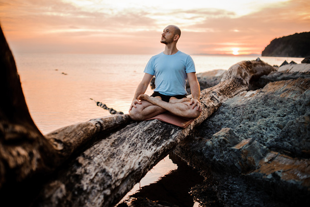 男は海の夕日近く壊れた木の瞑想します。屋外ヨガ - 写真・画像