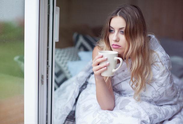 junge schöne blonde Frau mit einer Tasse Kaffee sitzt zu Hause auf dem Bett  - Foto, Bild