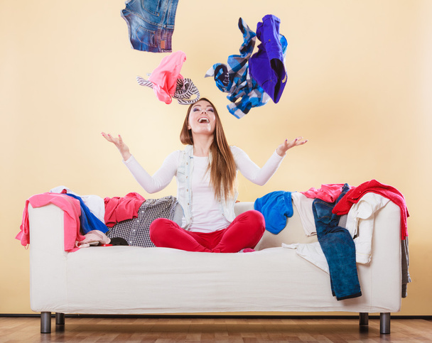 Happy woman on sofa in messy room throwing clothes - 写真・画像