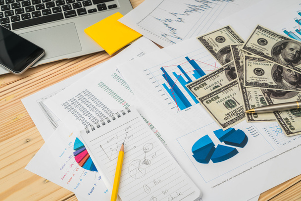 Фінансові графіки на столі з ноутбуком, телефоном, доларовим банком
 - Фото, зображення